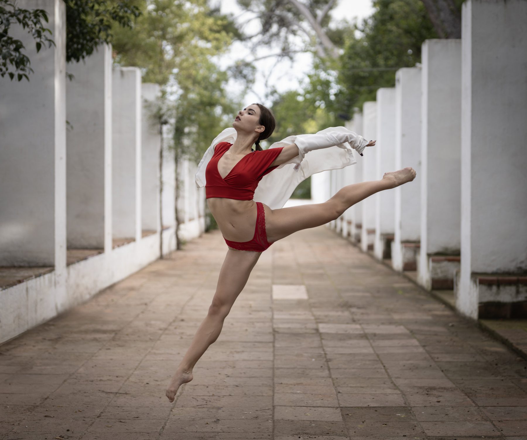 Ballerina jumping with extended leg and red dress in an outdoor corridor with white columns – artistic photography by VicmanArt