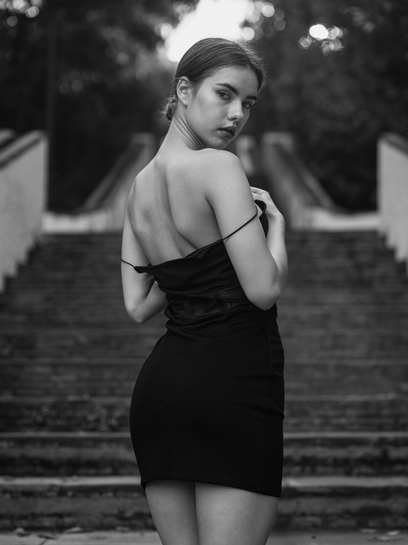 Model in a black dress from behind looking over her shoulder on park steps, black and white – artistic photography by VicmanArt