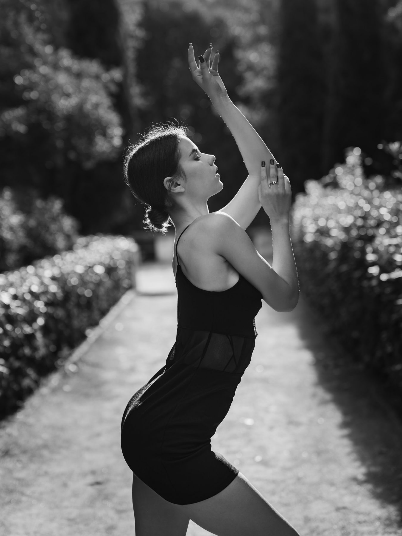 Ballerina in a black dress in profile raising her arms in a garden with cypresses, black and white – artistic photography by VicmanArt