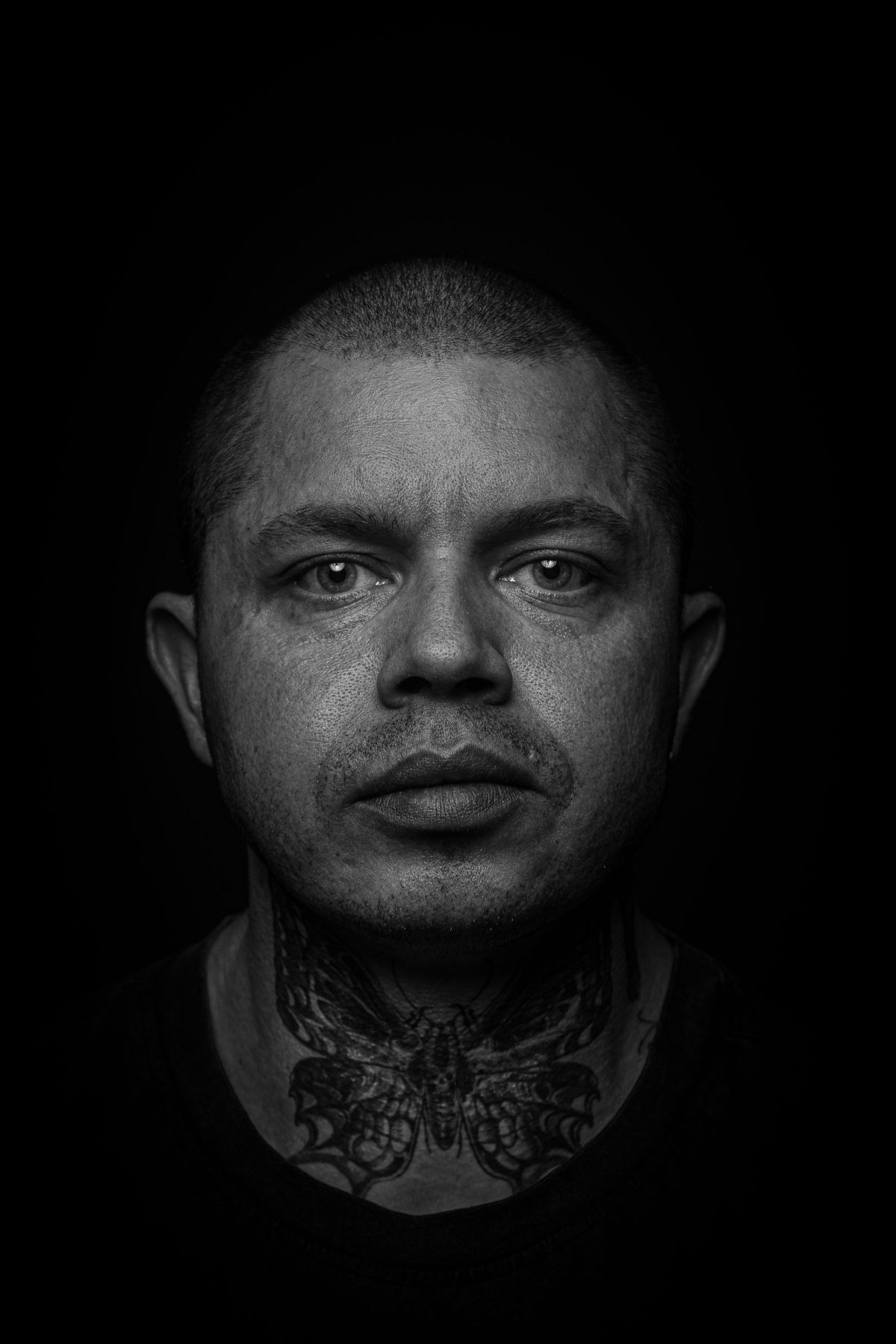Hombre con tatuaje en el cuello mirando a cámara con cabeza rapada, blanco y negro – fotografía de retrato editorial por VicmanArt