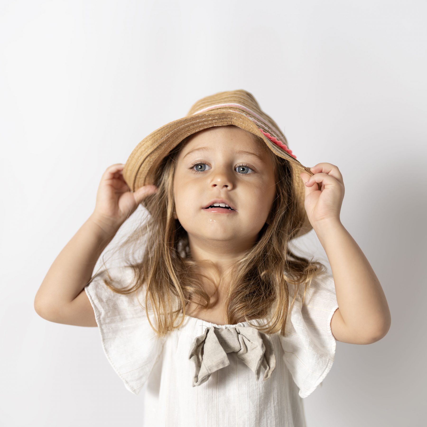 Niña pequeña con sombrero de paja y vestido blanco mirando a cámara, fondo gris – fotografía infantil por VicmanArt
