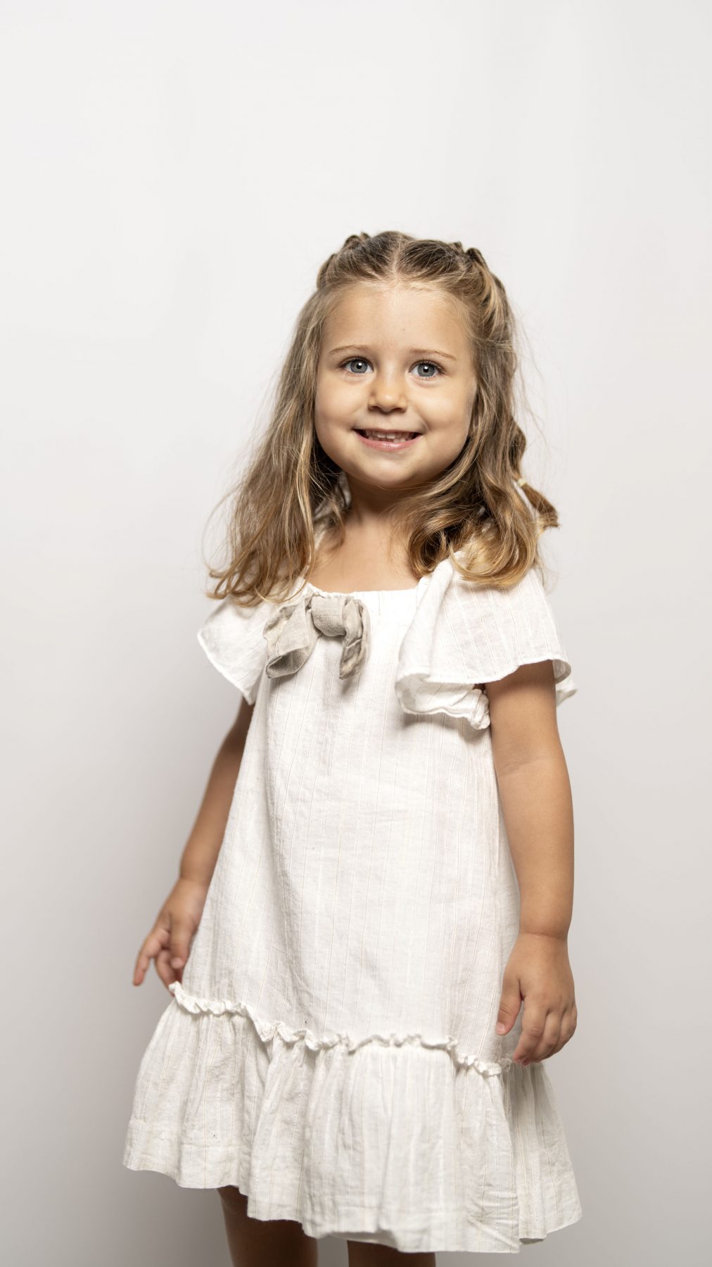 Niña pequeña con vestido blanco y lazos en el pelo sonriendo en estudio – fotografía infantil por VicmanArt