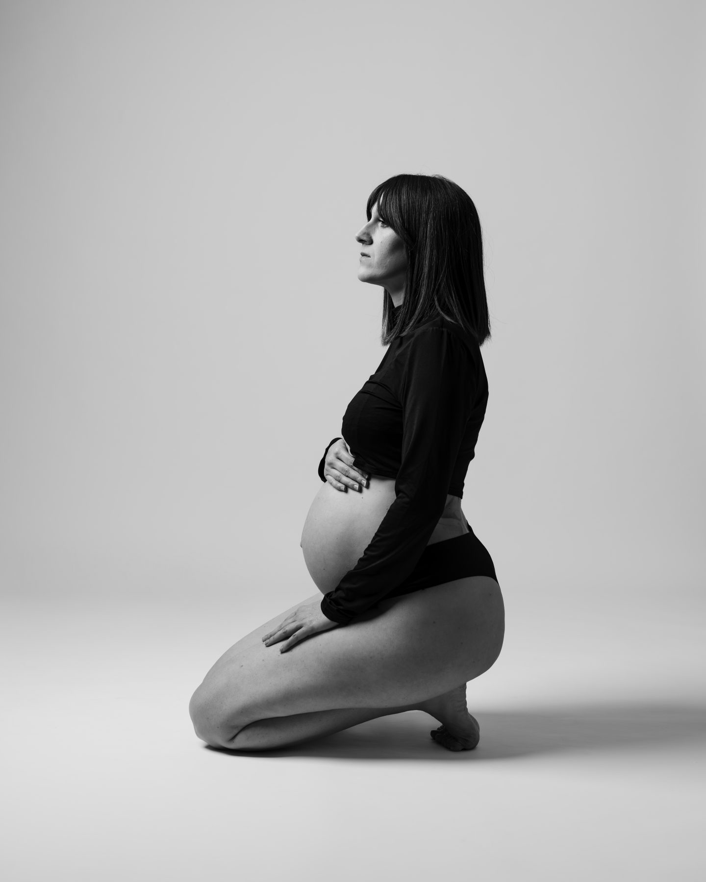 Mujer embarazada arrodillada de perfil en estudio minimalista, blanco y negro – fotografía de maternidad por VicmanArt.