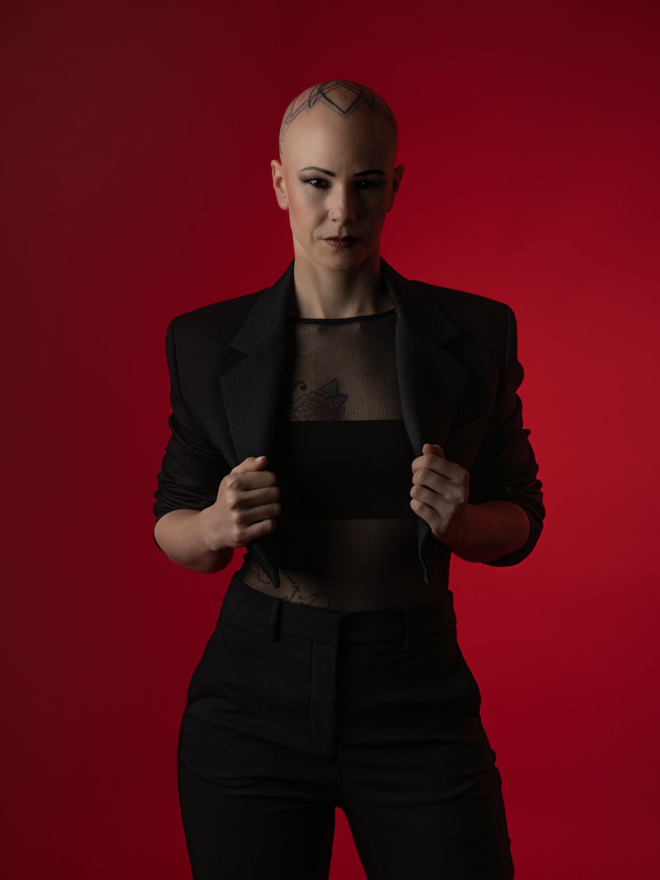Modelo con cabeza rapada y tatuajes en traje negro agarrando las solapas, fondo rojo – fotografía editorial de moda por VicmanArt.