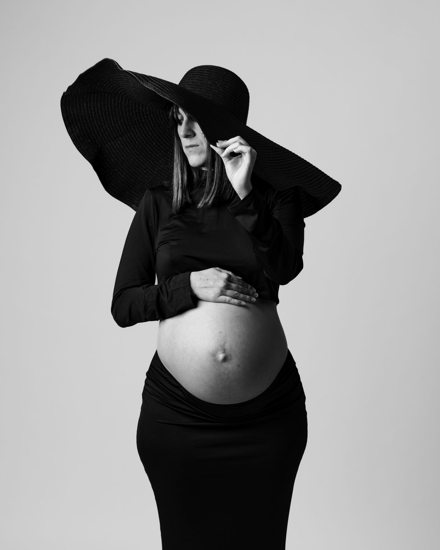 Mujer embarazada sosteniéndose el sombrero negro de ala ancha, estudio en blanco y negro – fotografía de maternidad por VicmanArt.