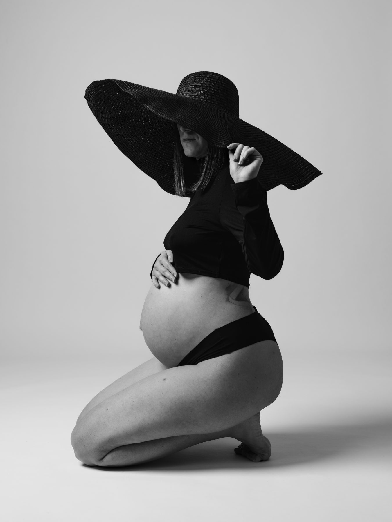 Mujer embarazada arrodillada con gran sombrero negro, estudio en blanco y negro – fotografía de maternidad por VicmanArt.