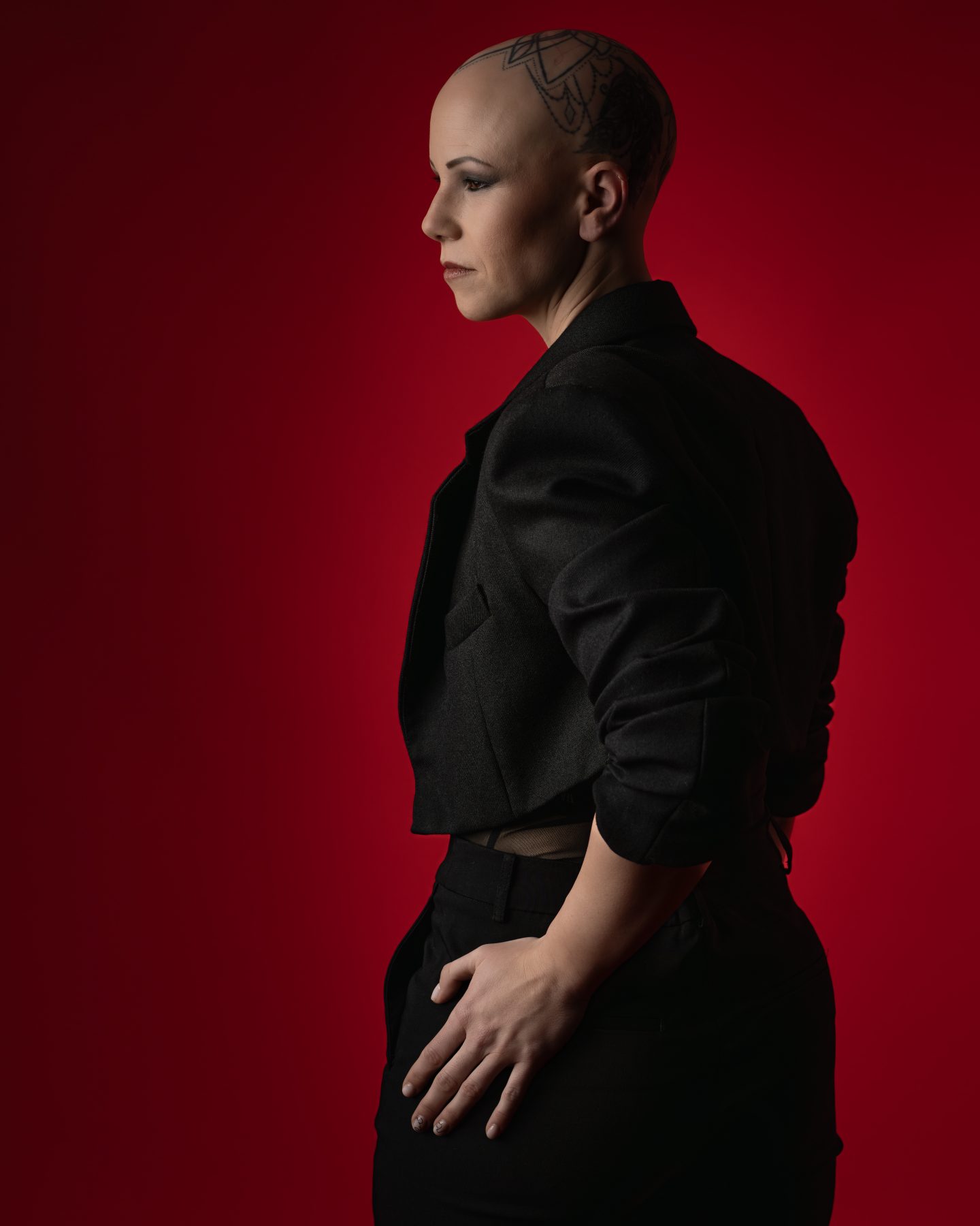 Modelo con cabeza rapada de perfil con blazer negro recortado, fondo rojo – fotografía editorial de moda por VicmanArt