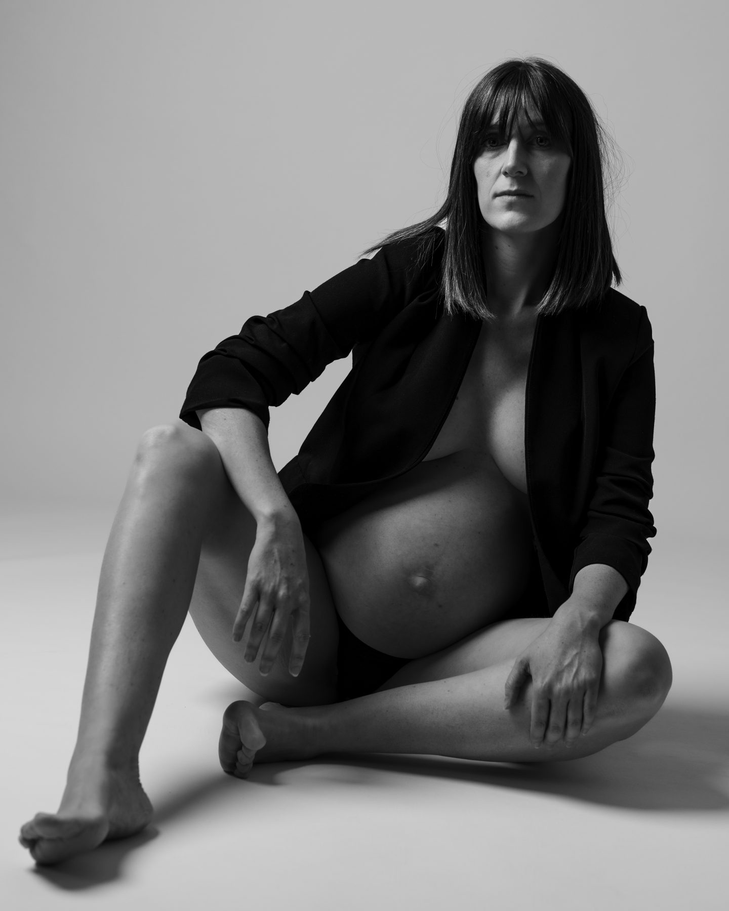 Mujer embarazada sentada en el suelo con blazer negro abierto, estudio en blanco y negro – fotografía de maternidad por VicmanArt.
