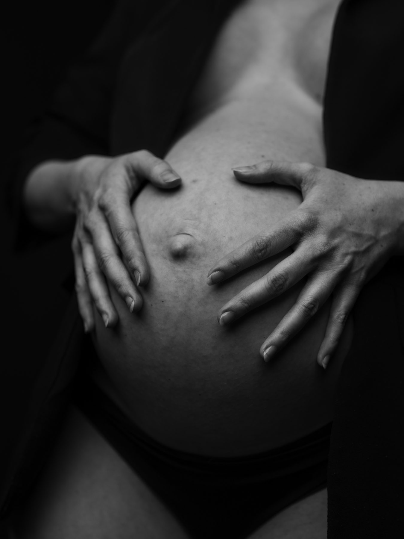 Primer plano de manos sobre barriga embarazada, fondo oscuro – fotografía de maternidad por VicmanArt.