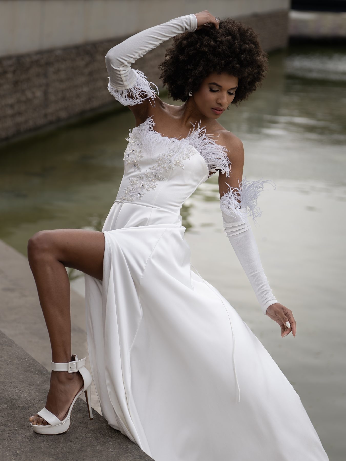 Model in a white feathered dress posing next to an urban fountain in Barcelona – editorial fashion photography by VicmanArt.