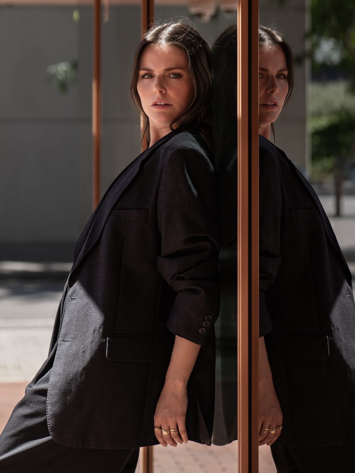 Modelo con traje gris apoyada en cristal, retrato de tres cuartos con anillos dorados visibles – fotografía editorial de moda por VicmanArt.