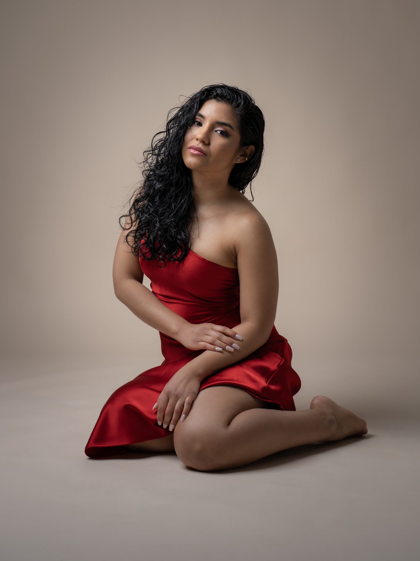 Model in a satin red dress sitting on the studio floor, beige background – portrait photography by VicmanArt