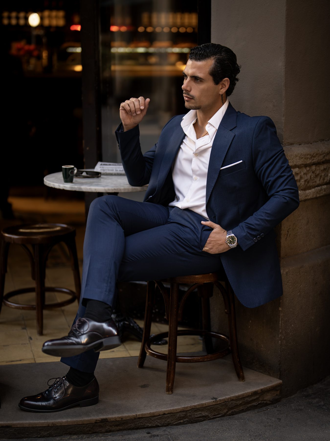 Modelo masculino con traje azul marino sentado en taburete en una terraza de Barcelona, cuerpo entero – fotografía editorial de moda por VicmanArt.