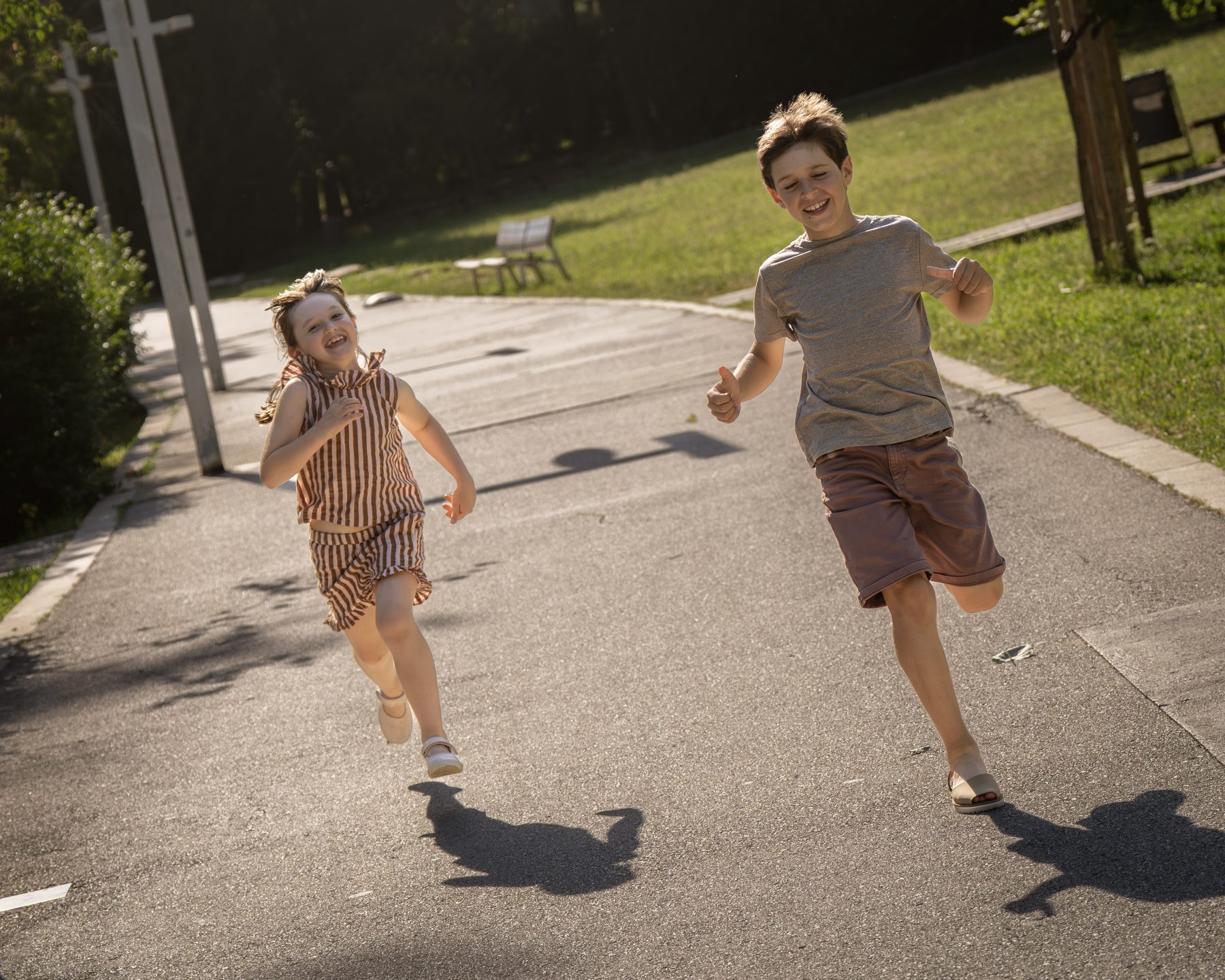 Dos hermanos corriendo y riendo en el paseo de un parque de Barcelona al sol – fotografía de familia por VicmanArt