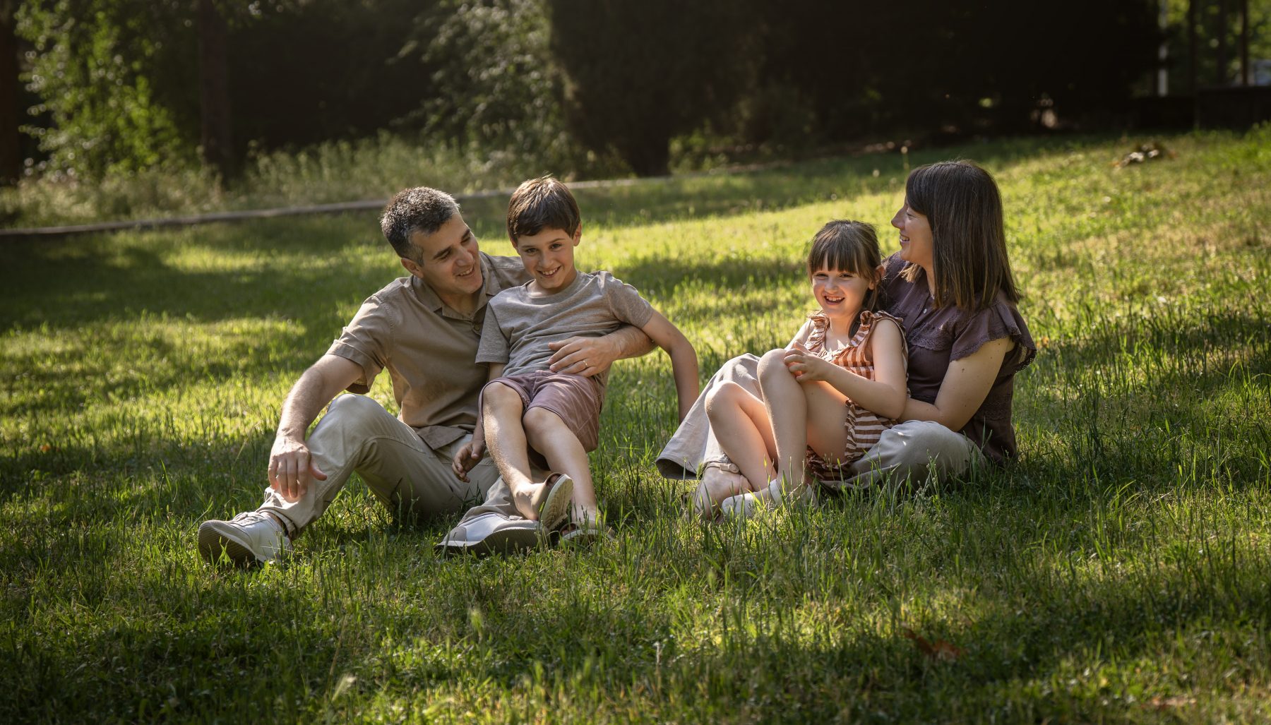 Familia de cuatro miembros riéndose juntos sentados en la hierba al atardecer, parque de Barcelona – fotografía de familia por VicmanArt